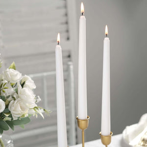 Three white taper candles lit in elegant gold holders next to a floral arrangement.