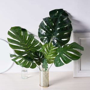 Artificial monstera leaves in a gold vase on a light wood surface.
