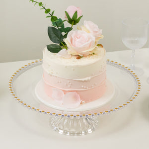Three-tiered wedding cake decorated with roses and pearls on a glass pedestal.
