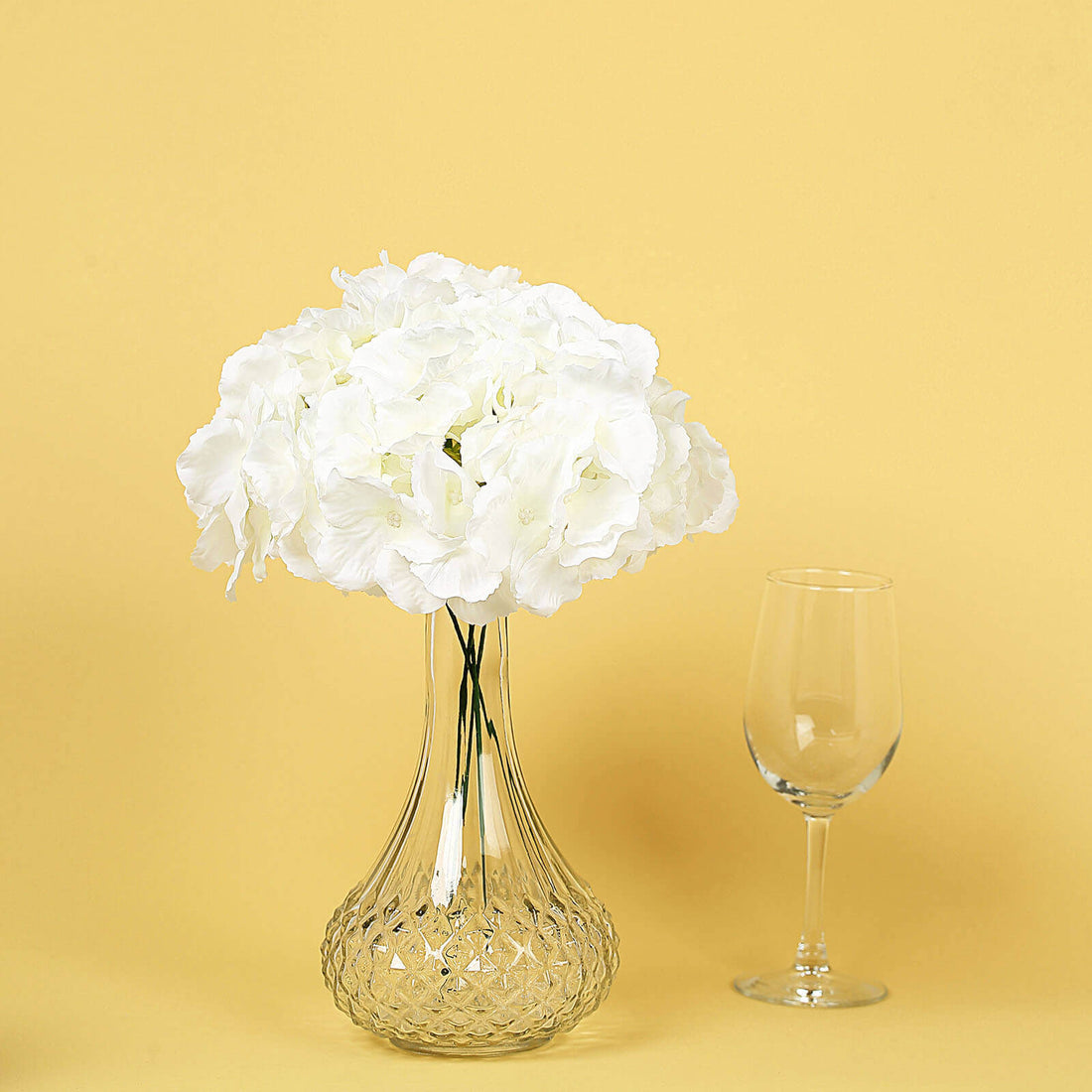 White hydrangea flowers in a clear glass vase with a wine glass on a yellow background.