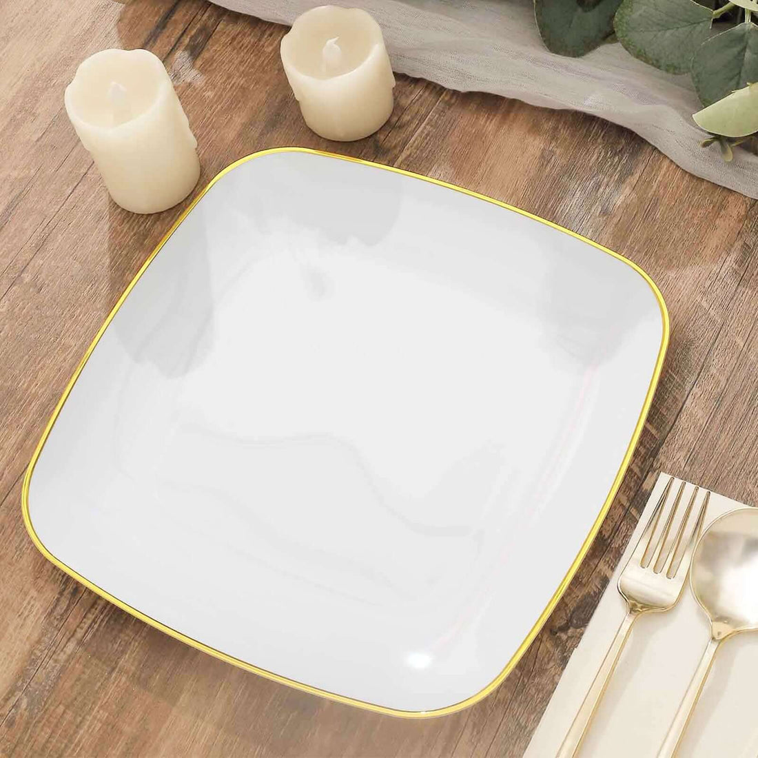 White square plate with gold rim on wooden table with candles and cutlery.