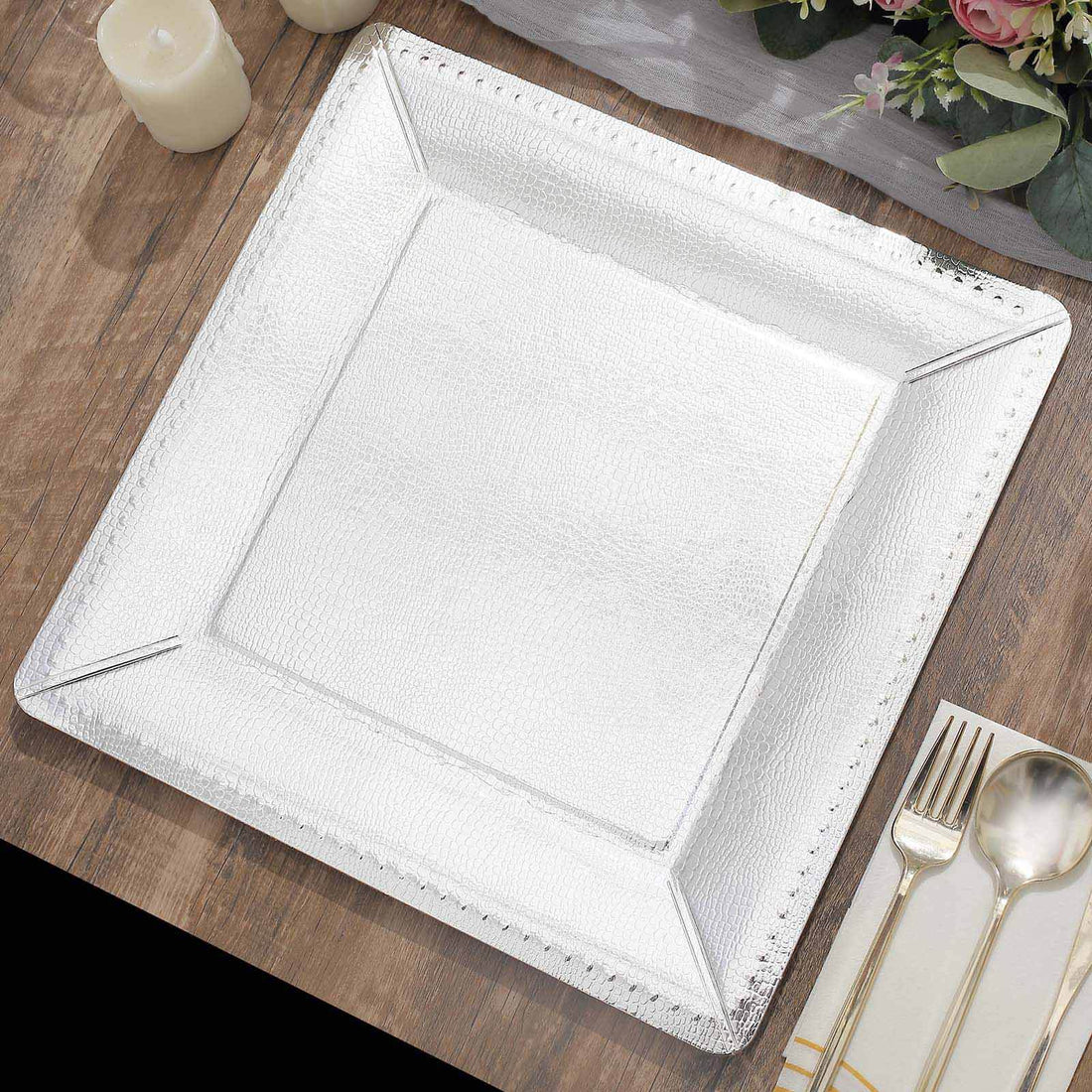 Silver square charger plate on a wooden table setting with candles and flowers.