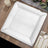Silver square charger plate on a wooden table setting with candles and flowers.