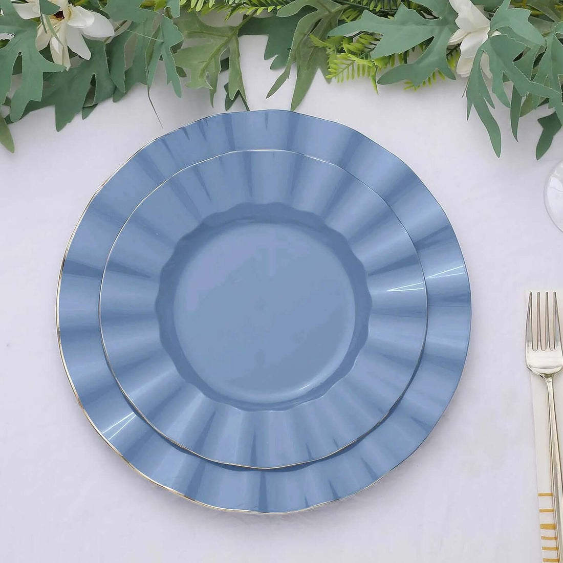 Blue decorative plates layered on a table with greenery and cutlery.