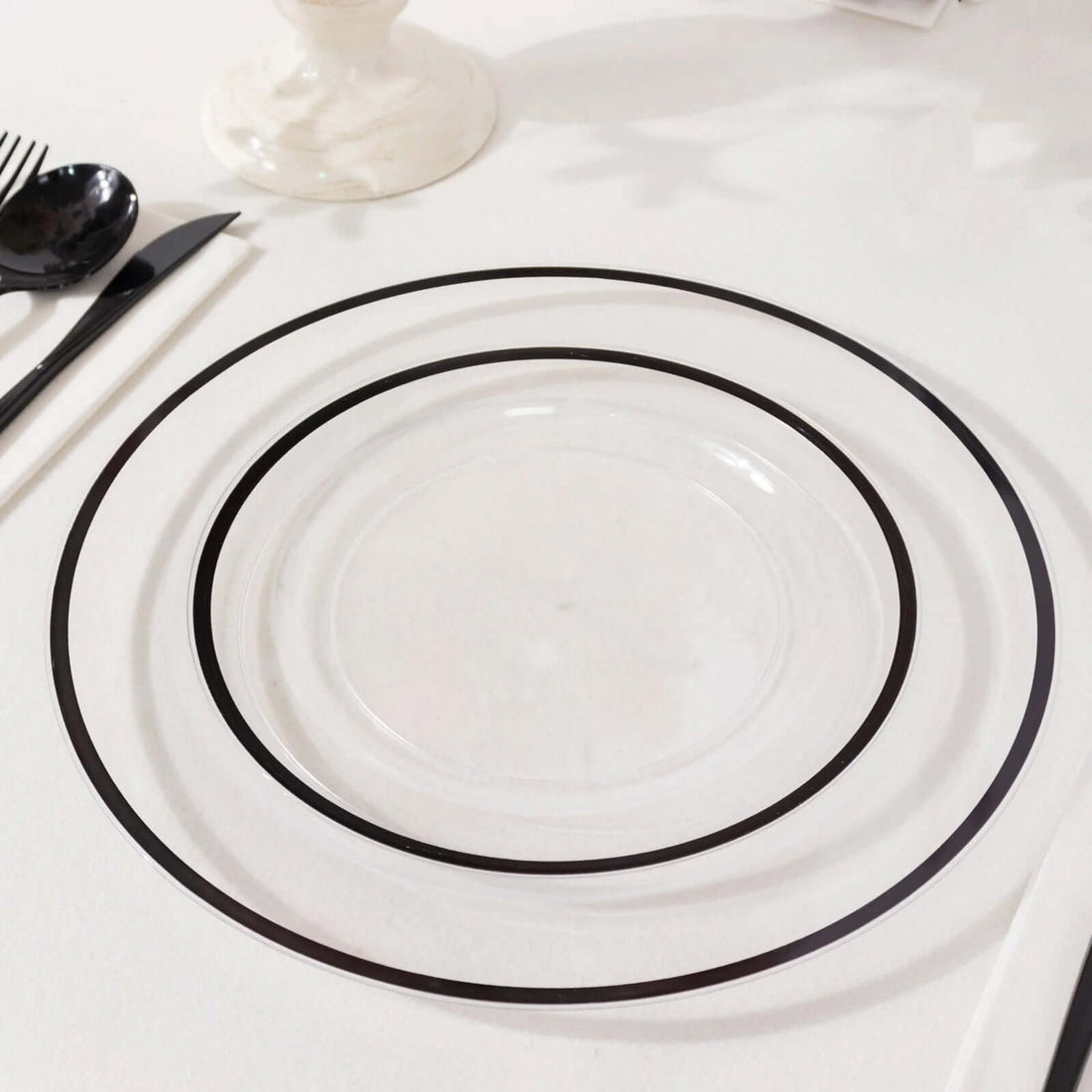 Clear glass plates with black rims in three sizes on a white table setting.