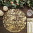 Gold textured round placemat on a wooden table with candles and greenery.