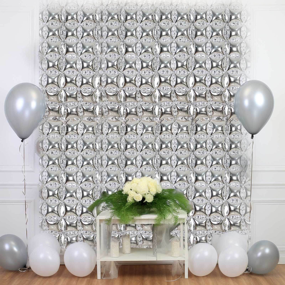 Party backdrop with silver balloons and white flowers on a table.