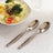 Stainless steel serving spoons next to a fresh salad on a table.
