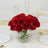 Red roses in a crystal vase on a dining table with white plates and gold utensils.