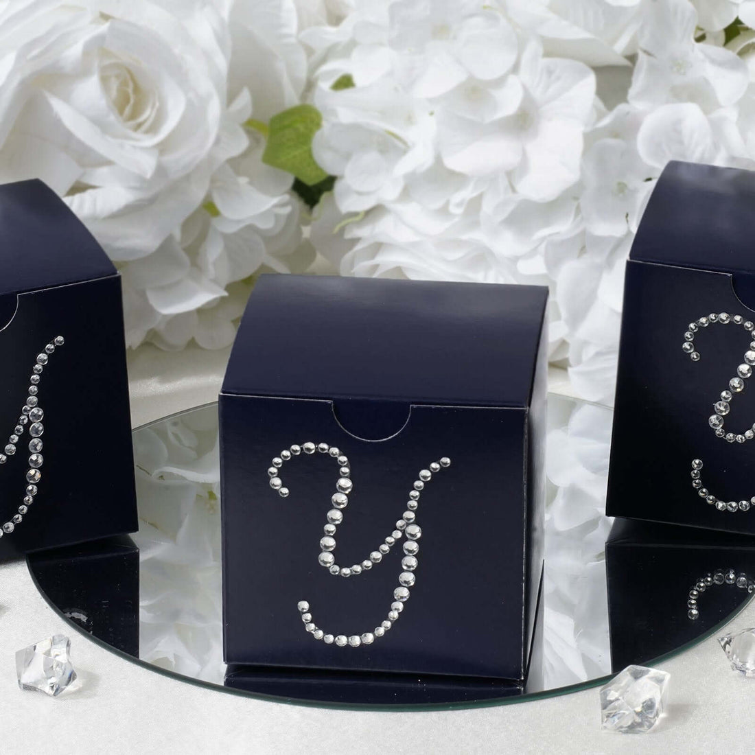 Navy gift boxes with rhinestone 'Y' on a mirrored surface surrounded by flowers. #whtbkgd