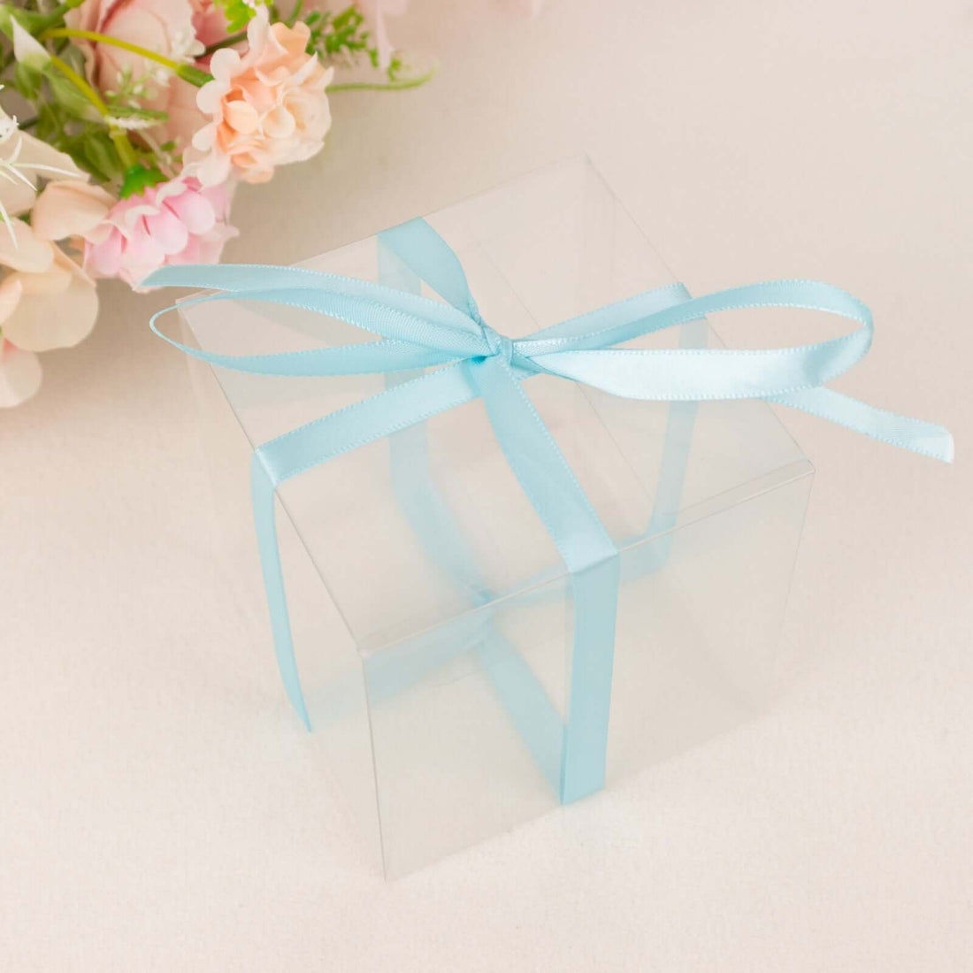 Clear gift box tied with a blue ribbon, surrounded by flowers.