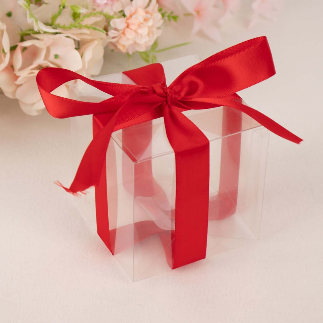 Clear gift box with a red ribbon and bow, surrounded by soft floral decor.