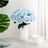Light blue artificial rose bouquet in a textured white vase with a cup beside it.