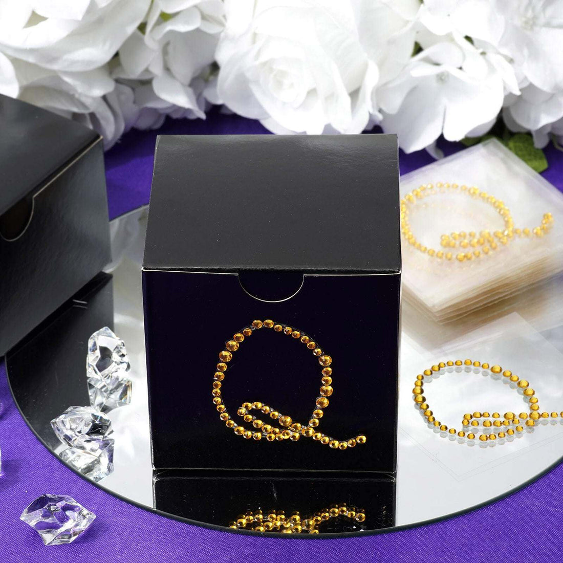 Black gift box with gold rhinestone letter "Q" on a mirrored surface.