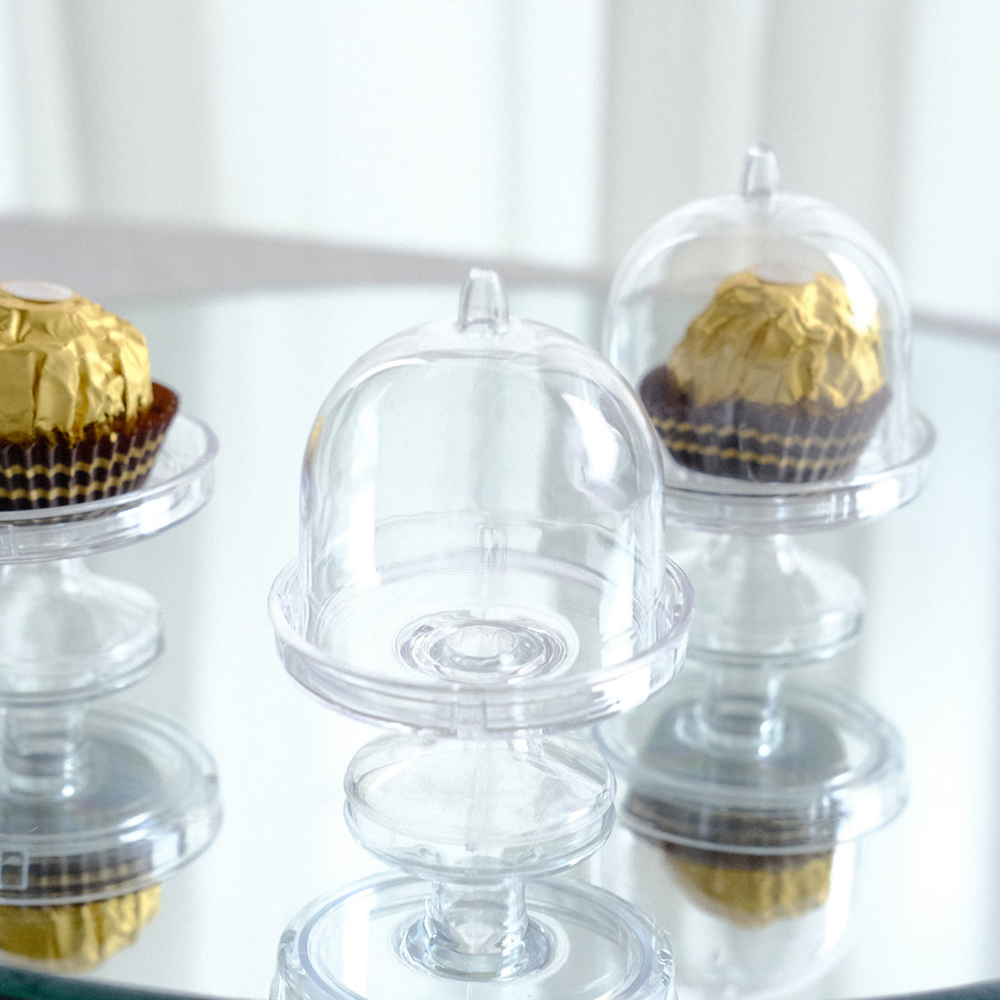 Clear cupcake cloches with gold-topped treats on a reflective surface.