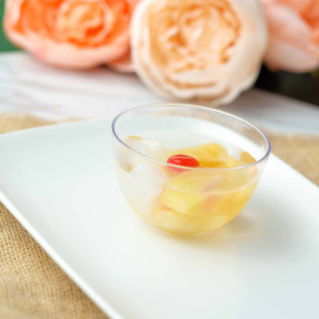 Fruit cocktail in a clear bowl on a white plate, with pink flowers in the background.
