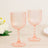 Two pink ribbed glass goblets on a wooden table with flowers in the background.