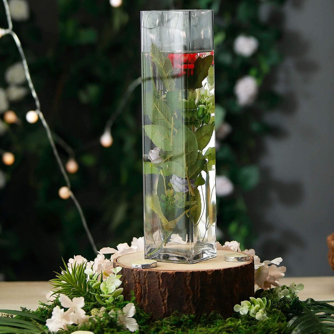 Clear glass vase filled with greenery and flowers on a wooden base.