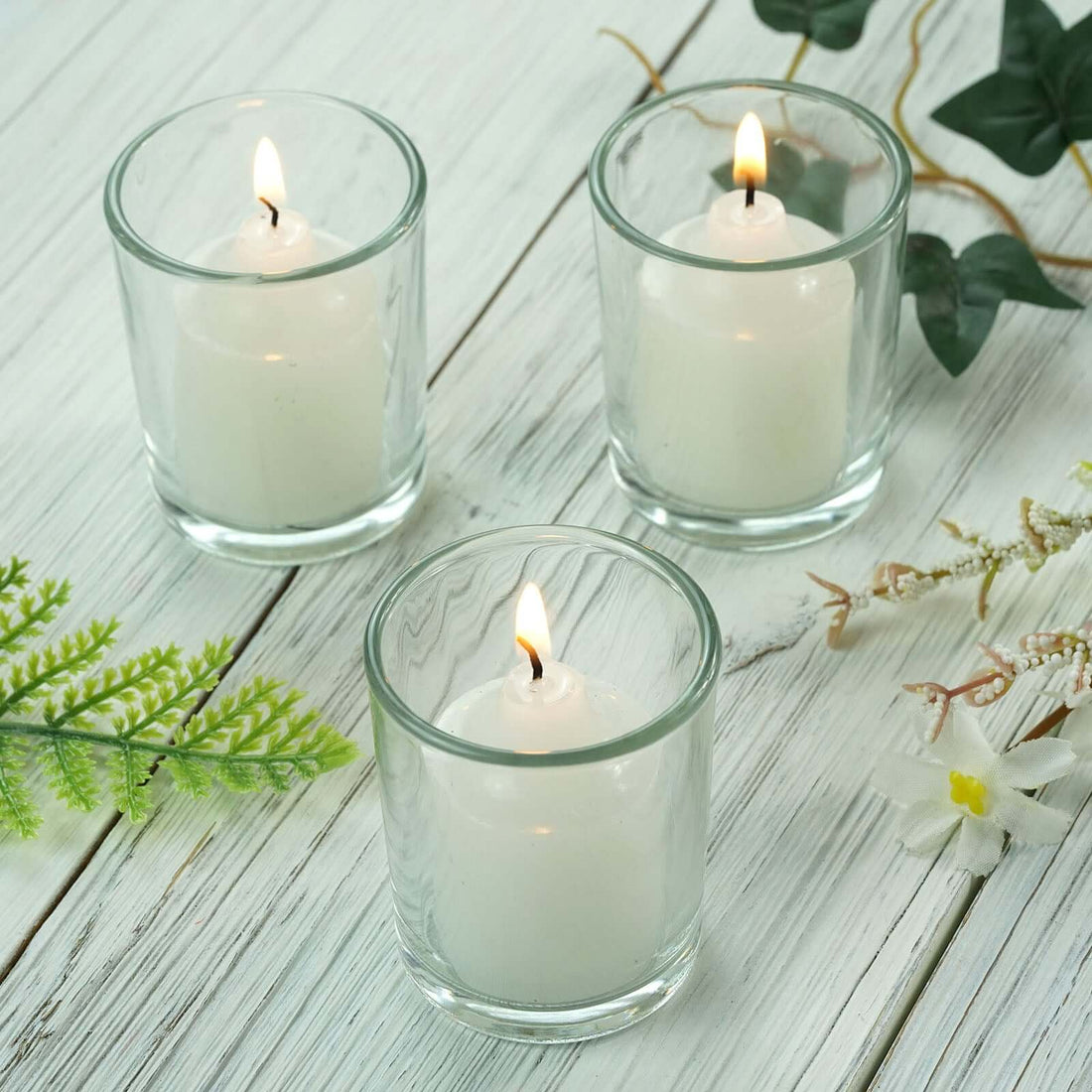 Set of three glass candles with lit wicks on a rustic wooden surface.