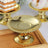 Gold cake stand on a wooden tray with desserts in the background.