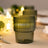 Green ribbed glass candle holder with warm glowing lights in the background.