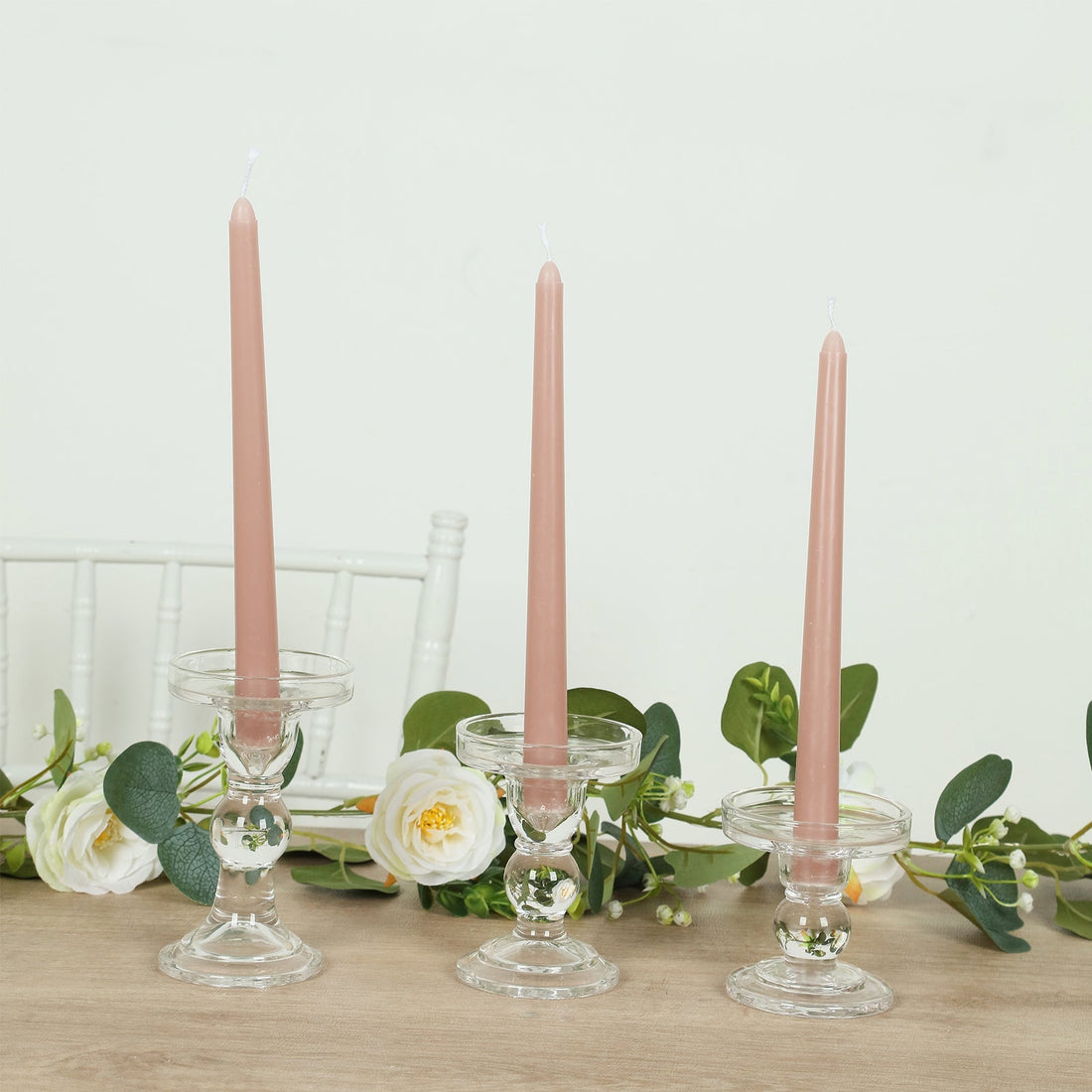 Pink taper candles in glass holders with greenery and flowers on a table.