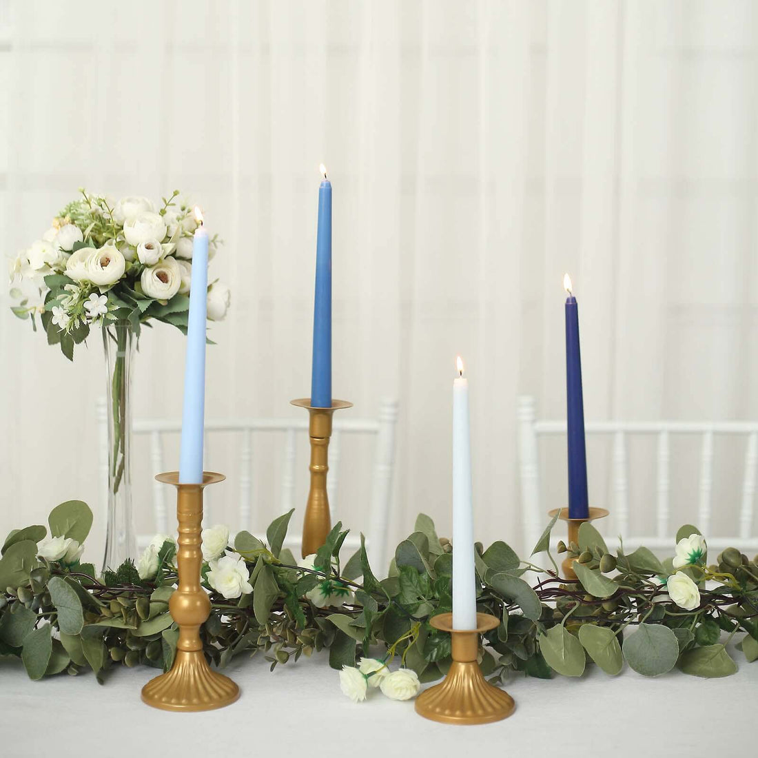 Elegant candle display with blue and gold accents, surrounded by greenery and flowers.