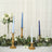 Elegant candle display with blue and gold accents, surrounded by greenery and flowers.