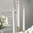 Three white taper candles lit in elegant gold holders next to a floral arrangement.