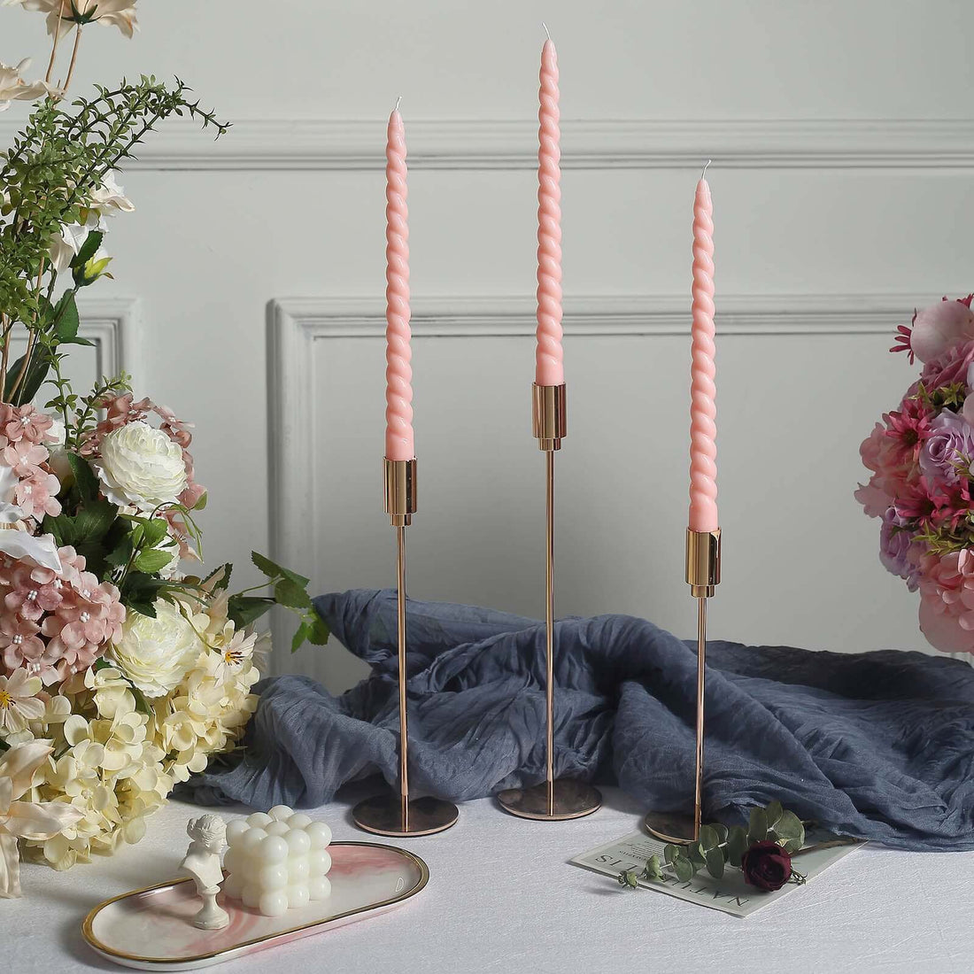 Elegant pink twisted candles in gold holders on a styled table with flowers and fabric.