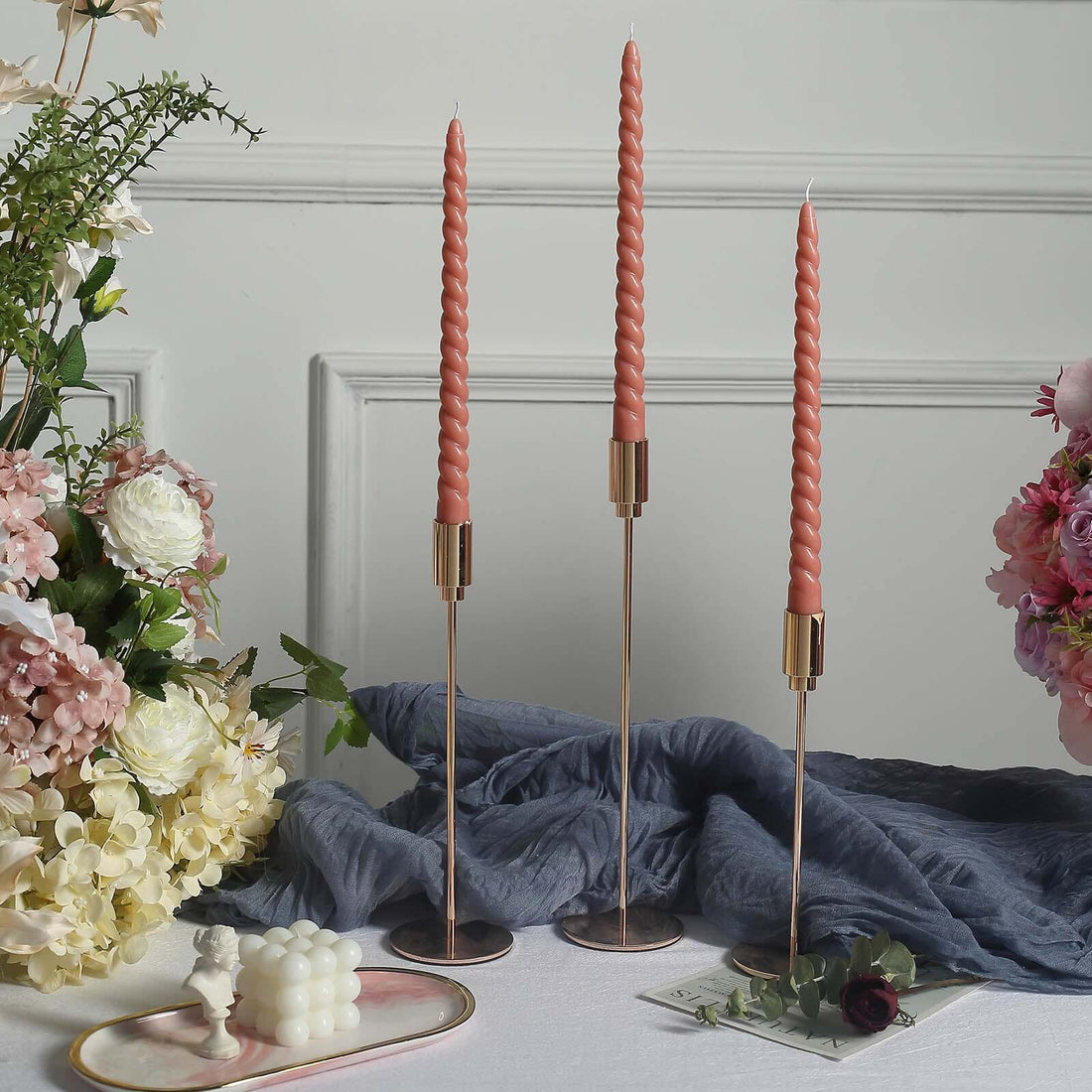 Elegant twisted candles in rose gold holders with floral arrangement backdrop.