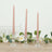 Three twisted pink candles in clear glass holders on a table with greenery and flowers.