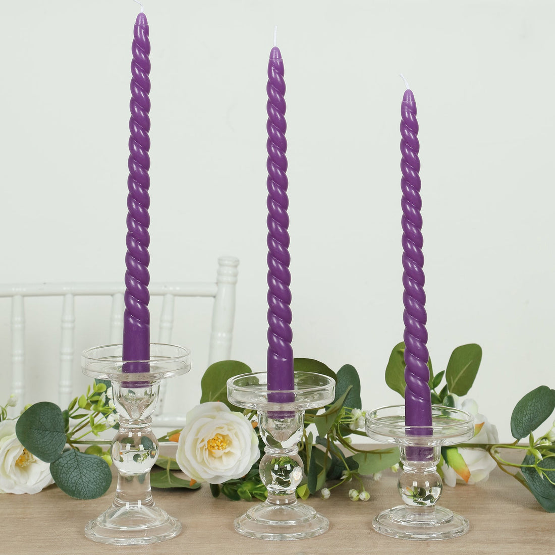Three purple twisted candles in clear glass holders on a neutral table setting.