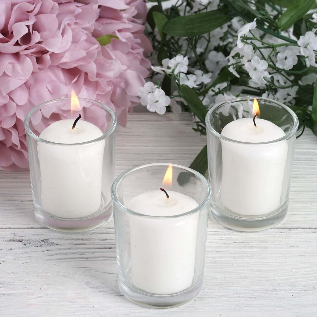 Three white candles in glass holders with pink flowers in the background.