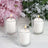 Three white candles in glass holders with pink flowers in the background.