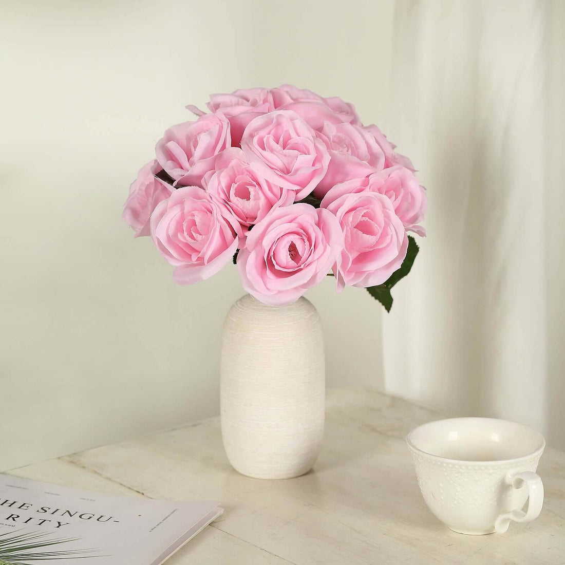 Pink rose bouquet in a white vase on a table with a cup and book.