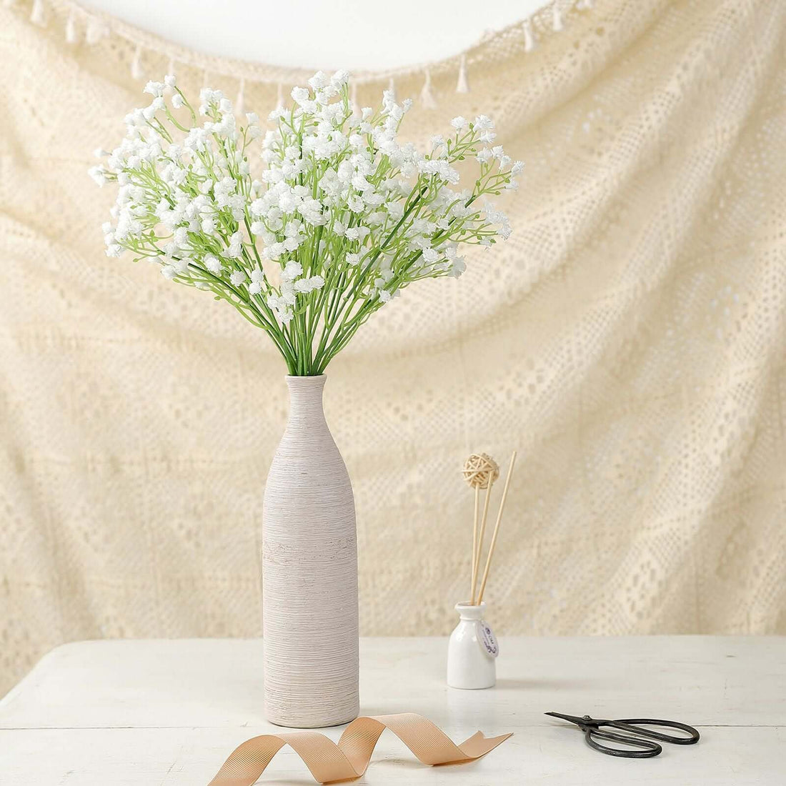 White flower arrangement in a textured vase on a table with decorative backdrop.