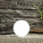 Glowing round outdoor lamp on gravel with stone wall backdrop.
