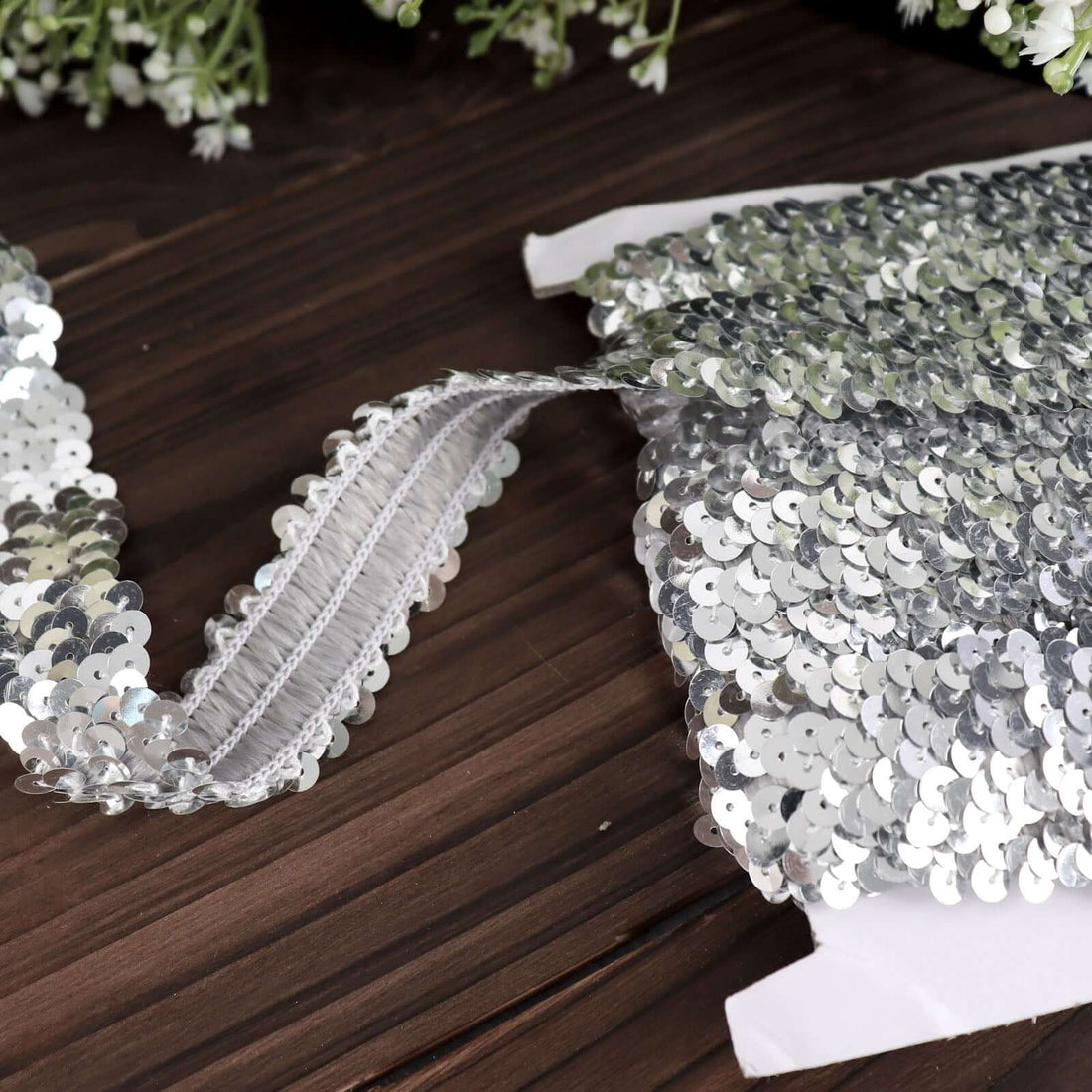 Silver sequin trim on a wooden surface with greenery in the background.