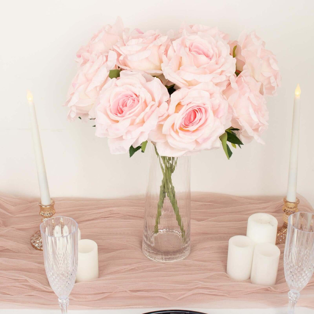 Pink artificial roses in a clear vase with candles on a soft pink table runner.