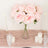Pink artificial roses in a clear vase with candles on a soft pink table runner.