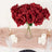 Red artificial roses in a glass vase on a table with candles.