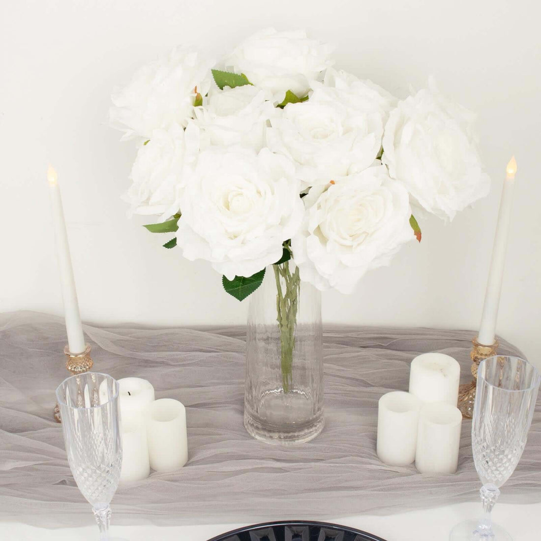 White roses in a glass vase with candles on a table setting.