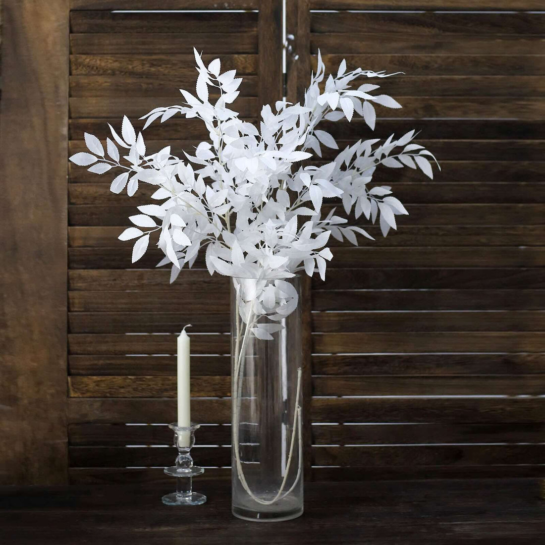 White faux leaf arrangement in a clear vase with candle on a wooden surface.