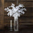White faux leaf arrangement in a clear vase with candle on a wooden surface.