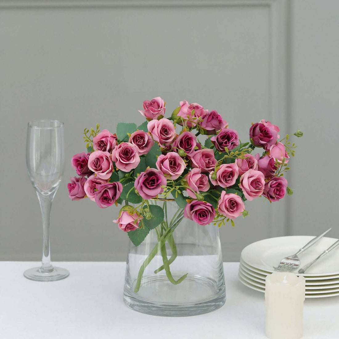 Pink roses in a clear vase on a table with dinnerware and a glass