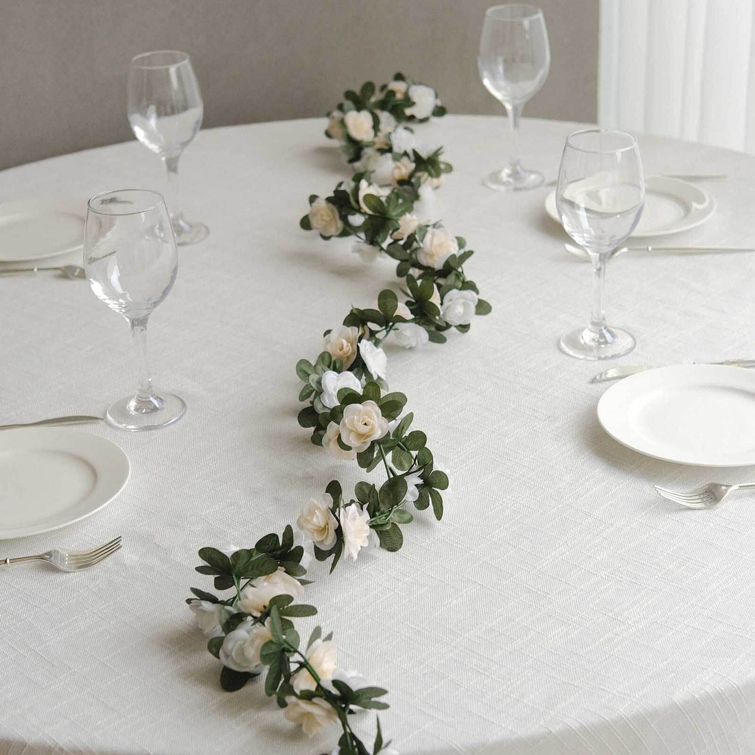 Floral table runner with white roses and greenery on a dining table setting.