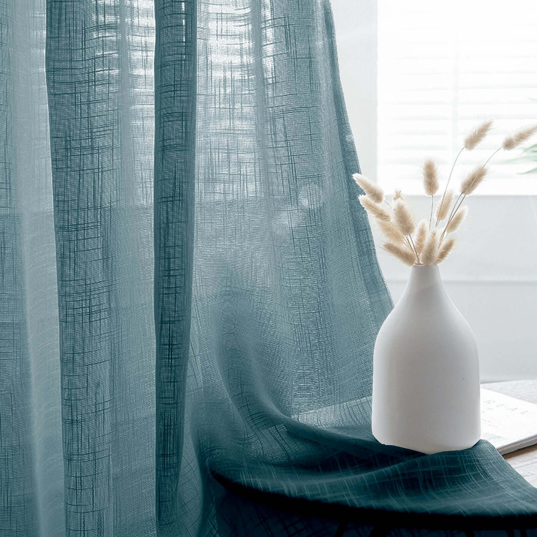 Sheer teal curtain draping elegantly beside a minimalist vase and dried flowers.