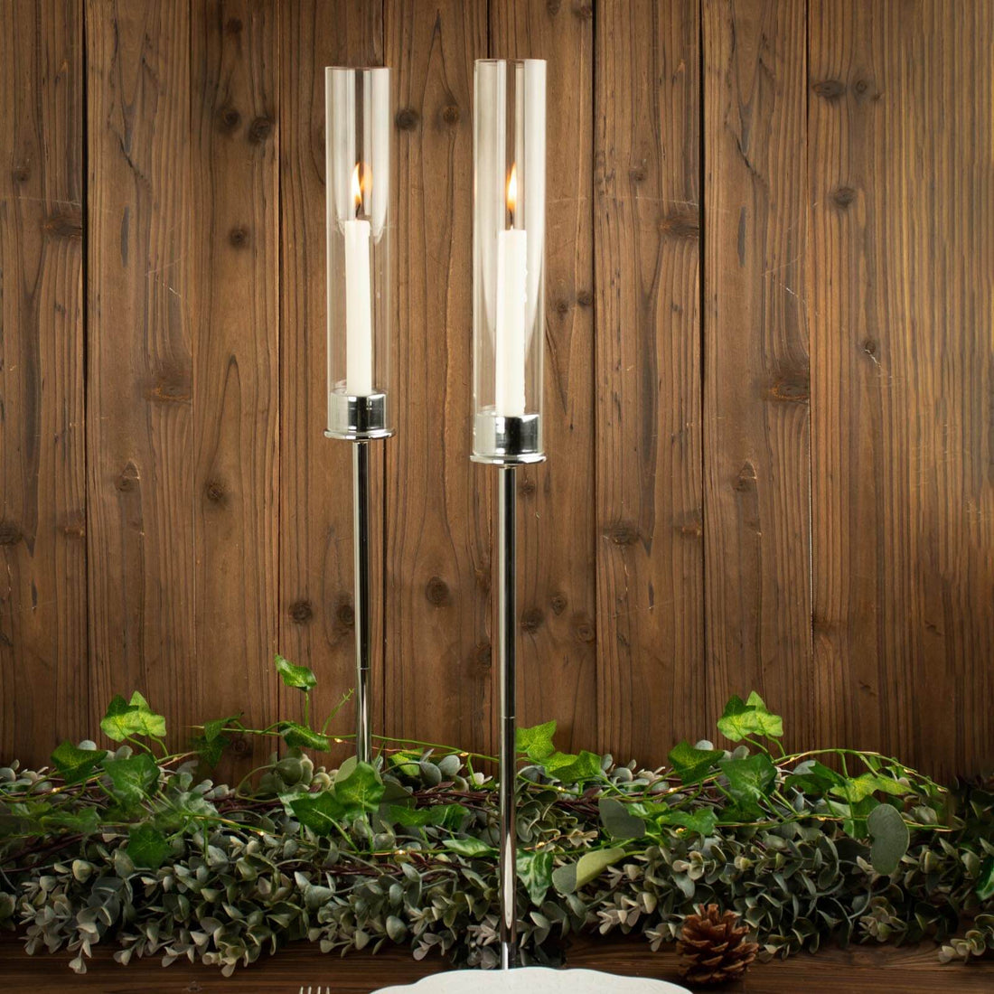 Elegant candle holders with glass cylinders and chrome bases, surrounded by greenery.