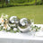 Decorative silver spheres on a table with flowers and glassware in a garden setting.